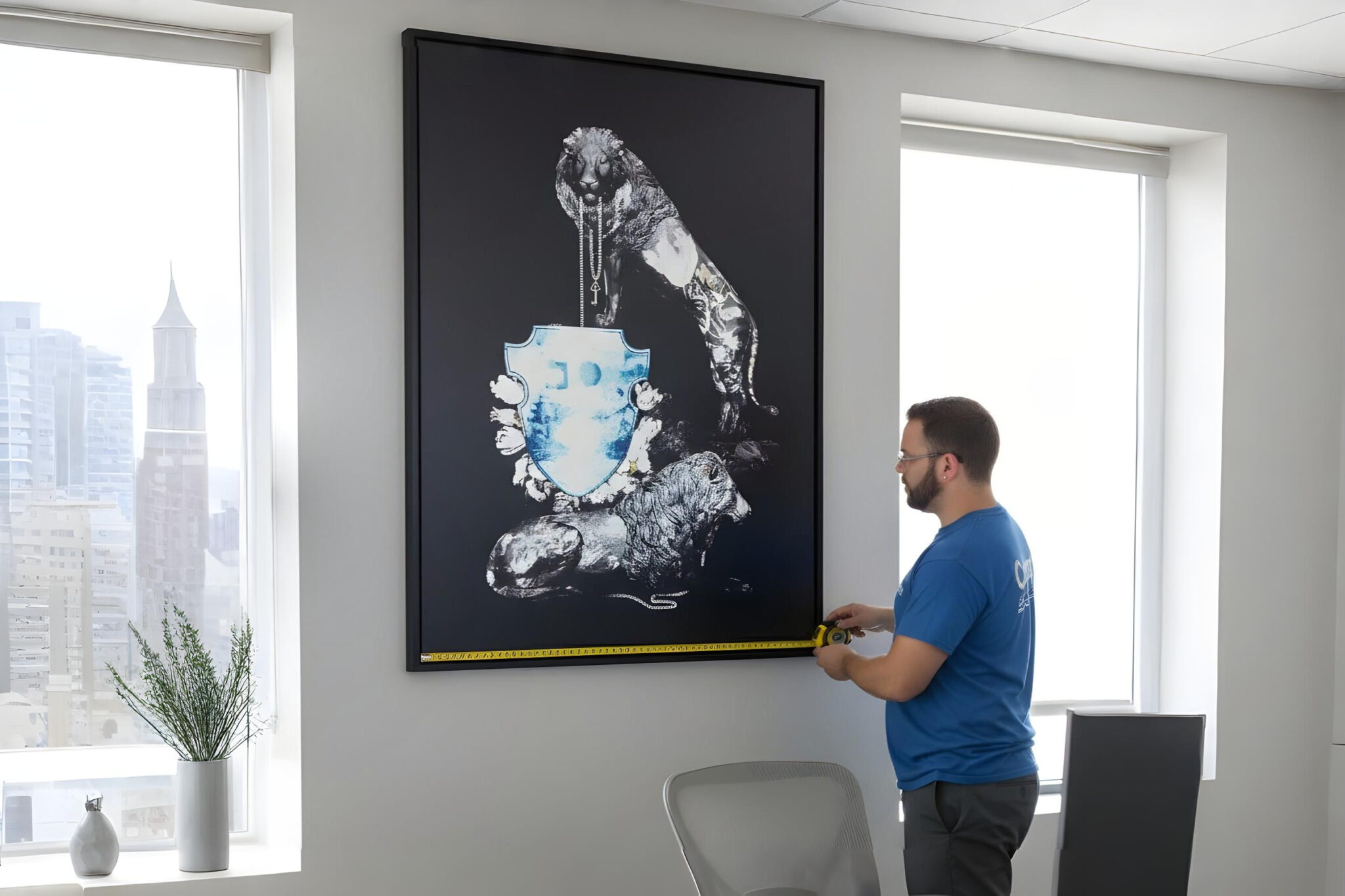 A professional mover wearing a blue t-shirt uses a yellow tape measure to gauge the width of a large, framed black-and-white artwork depicting a lion. The piece is mounted on a light grey office wall next to a large window, illustrating the initial inventory and planning phase where artwork is evaluated and measured.