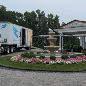 Clancy Relocation & Logistics truck parked at a grand home with a circular flowerbed and fountain, loading ramp to the covered porch and movers visible — moving services.