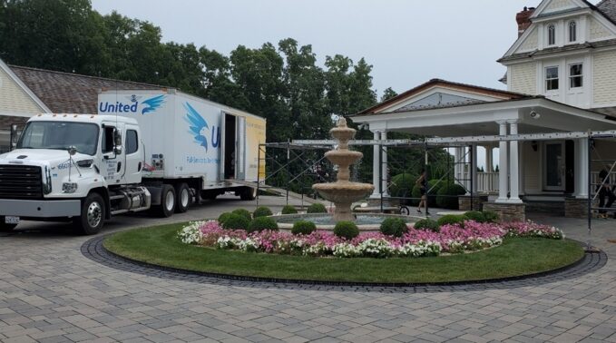 Clancy Relocation & Logistics truck parked at a grand home with a circular flowerbed and fountain, loading ramp to the covered porch and movers visible — moving services.