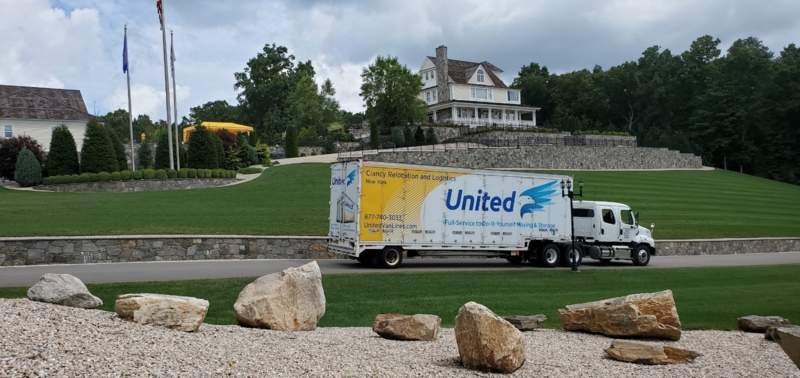 Clancy Relocation & Logistics truck parked on a driveway below a large hilltop house with landscaped lawn and stone walls — moving services.