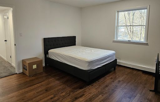 Minimalist bedroom with a wrapped headboard, mattress on a black platform bed, one cardboard box on hardwood floor — home movers.