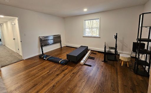 Empty living room with hardwood floors, wrapped headboard and furniture pieces, moving blanket and disassembled items staged for pickup — home movers.